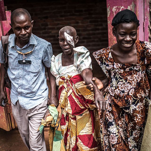 This photo shows an old Ugandan woman with an eye patch being helped by 2 people (a man and a woman)