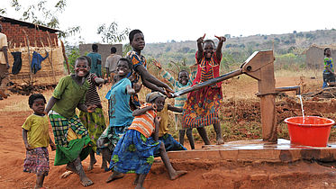 This photo shows a group of children at a water tap community point smiling and laughing together.