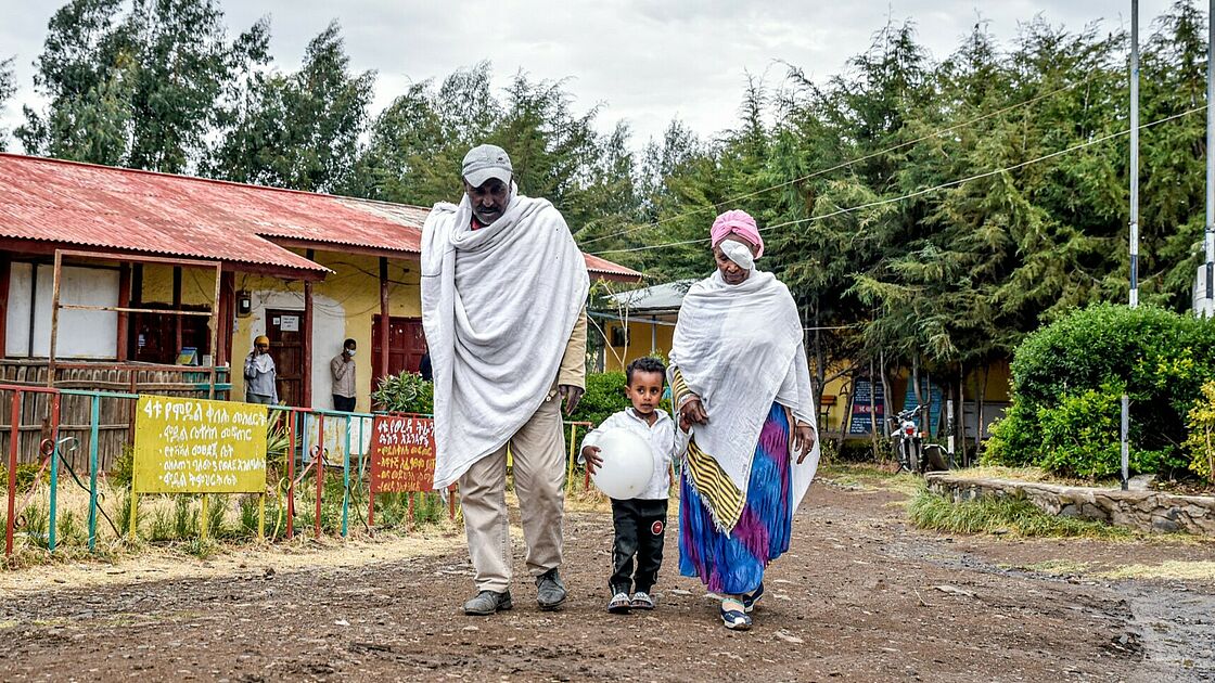 The image captures a moment of everyday life, featuring a family walking together along a dirt road. An older man and woman, both draped in traditional white garments with touches of colour, flank a young child holding a round white object, possibly a ballon. They are walking past a building with a red roof and colourful signage in what appears to be Amharic script, suggesting the setting is in Ethiopia or a region with similar language and customs. The surrounding environment has lush greenery, and it is a cloudy day. 