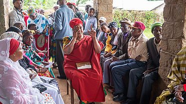 This photo showsa a group of Tanzanians in colourful traditional clothes sitting in a shed outside listening to someone give a speech.