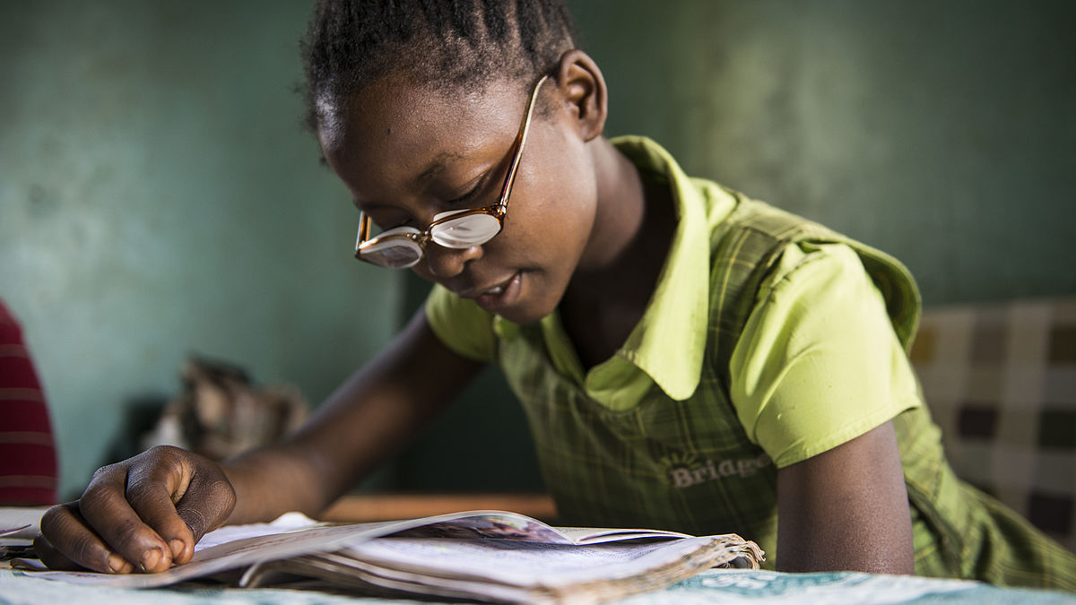Christine is sitting on a table at home reading a book. She is using her new pair of glasses given by CBM partner, Sabatia Eye Hospital in western Kenya.