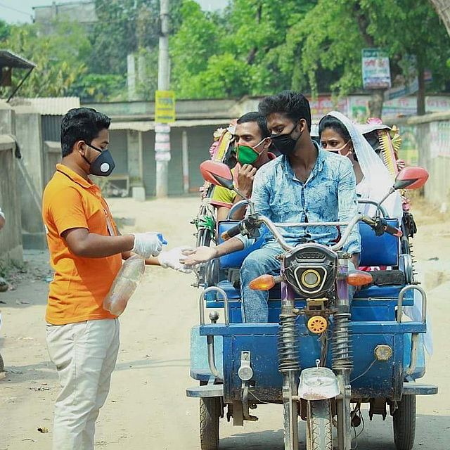 This photo shows a man spraying hand disinfectant on the hands of a rikshaw puller in Bangladesh. There are 2 women in the rikshaw. Everyone is wearing masks.