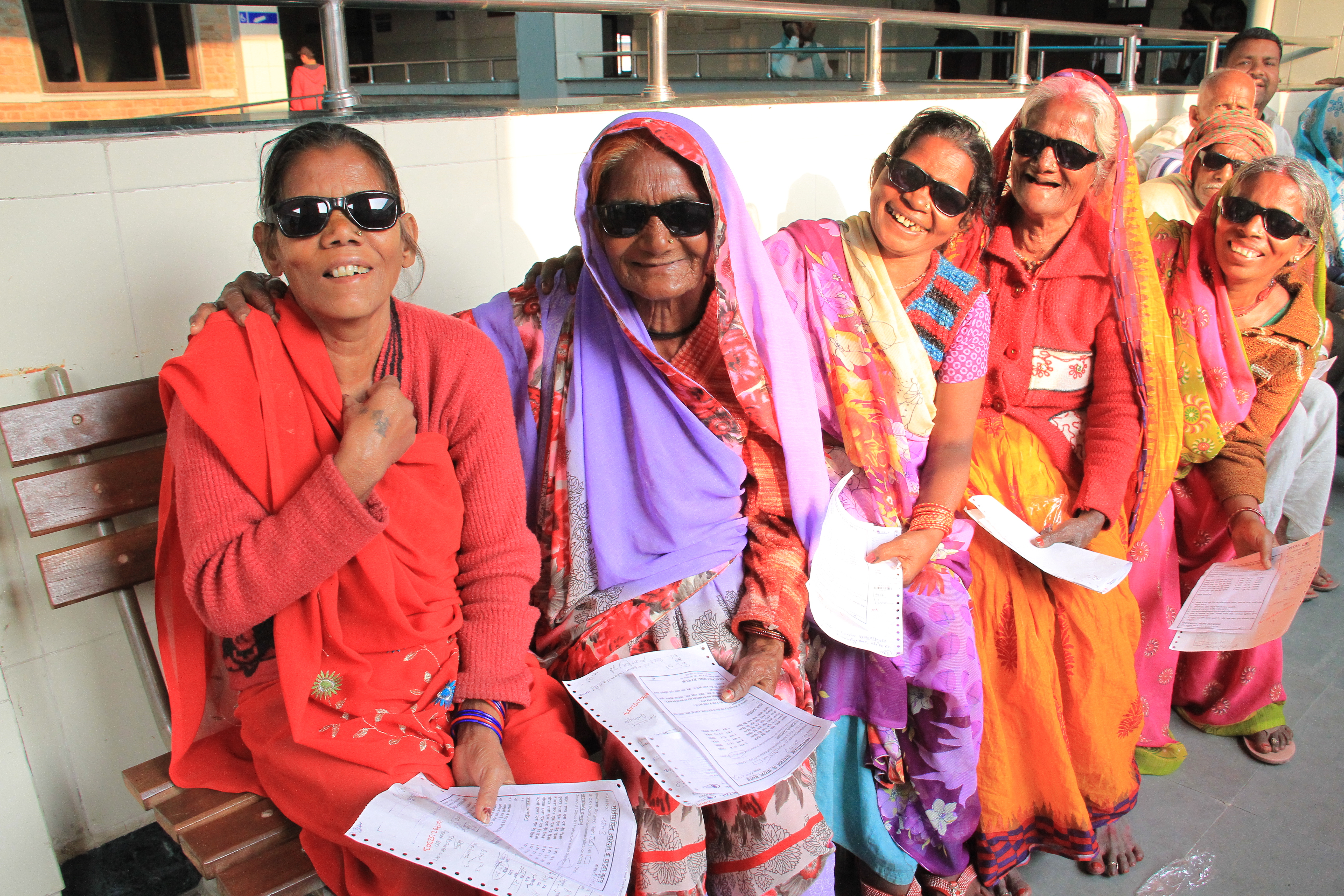 This image shows older women wearing colourful saris and sunglasses, sitting on a bench in a hospital and smiling at the camera.