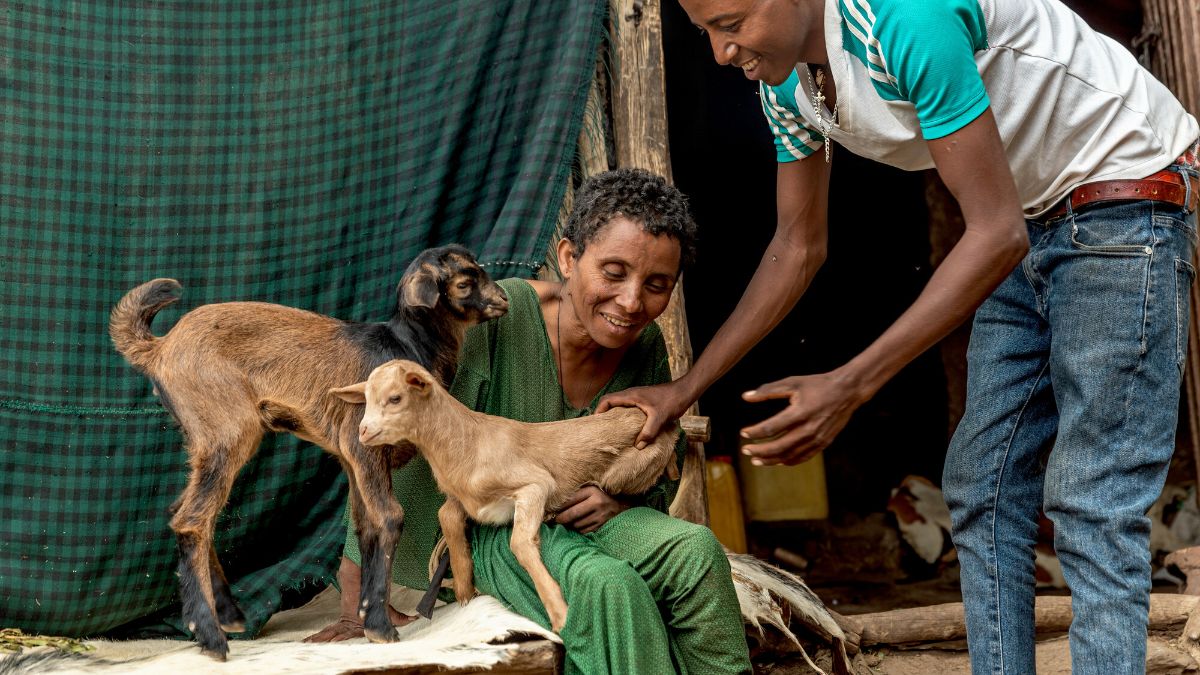 A woman sits and smiles while holding a pair of kid goats. A young man stands beside her, also smiling and offering assistance.