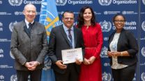 Four people standing in front of a backdrop featuring the World Health Organization (WHO) logo. One of the individuals at the centre holds a certificate that reads, "Certificate of Membership", awarded to the Christian Blind Mission (CBM). The people in the photo are dressed in formal attire. 