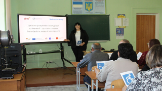 Presenter standing beside a projection screen, delivering a training session to participants in a classroom.