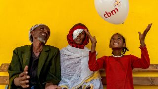 A man, a woman and a child sitting together. The child plays with a balloon, and the woman wears an eye patch over her left eye. 