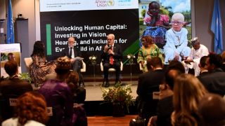 A group of people at a conference. Words on the banner behind the speakers read: Unlocking Human Capital. Investing in vision, hearing and inclusion for every child in Africa