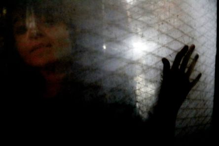 Eine Frau hinter einer Glasscheibe, auf der das Licht ein Rautenmuster zeichnet, lebt ihre Hand auf die Scheibe.