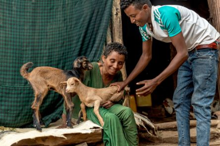 A woman sits and smiles while holding a pair of kid goats. A young man stands beside her, also smiling and offering assistance.