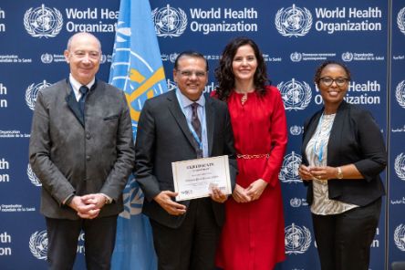 Four people standing in front of a backdrop featuring the World Health Organization (WHO) logo. One of the individuals at the centre holds a certificate that reads, "Certificate of Membership", awarded to the Christian Blind Mission (CBM). The people in the photo are dressed in formal attire. 