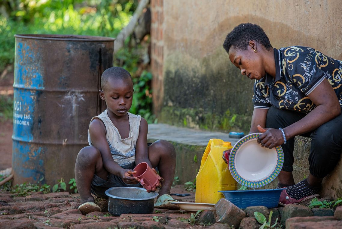 Ein Junge und eine Frau sitzen vor einer Hütte und spülen in zwei kleinen Schüsseln Geschirr.