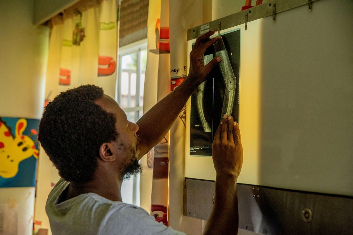 Ein Mann betrachtet ein Röntgenbild, das an einen Leuchtkasten geklemmt ist. Auf dem Bild sieht man weiß leuchtend zu einem O verbogene Beine.