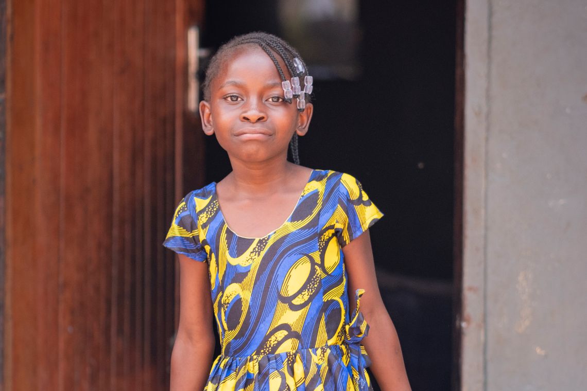 Ein etwa neunjähriges Mädchen steht in einem Türrahmen. In die kleinen Zöpfe seiner Frisur sind Glasperlen eingeflochten.