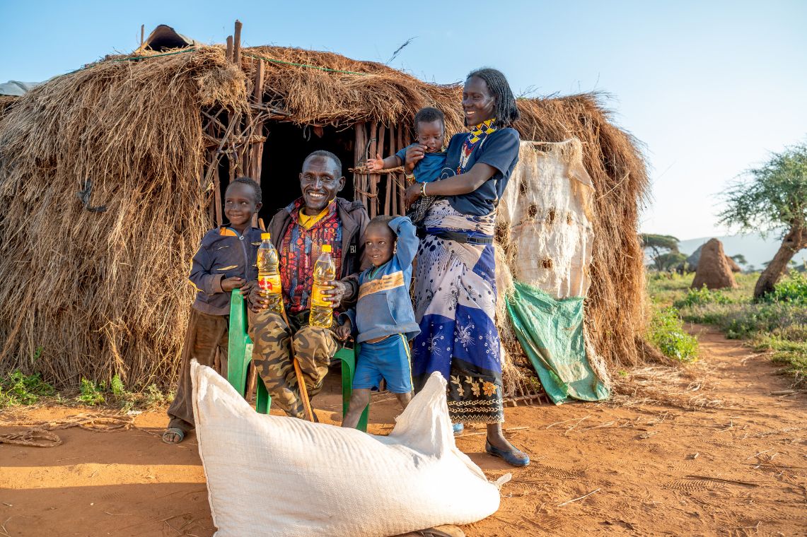 A smiling man is sitting in front of a thatched hut. To his left stands a small child; to his right stand a child and a woman holding a baby in her arms. In front of them lies a large, white sack.