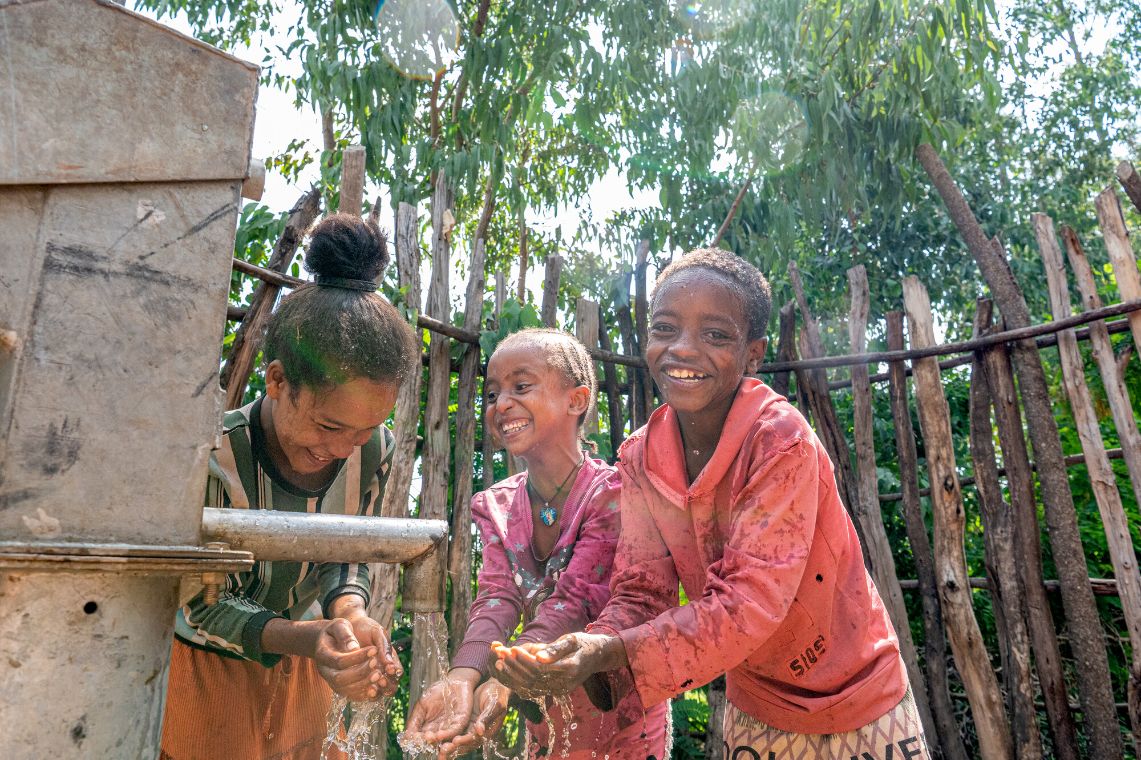 Three laughing children are standing by a pipe from which water is flowing, holding their hands under the stream.