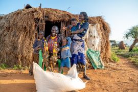 A smiling man is sitting in front of a thatched hut. To his left stands a small child; to his right stand a child and a woman holding a baby in her arms. In front of them lies a large, white sack.