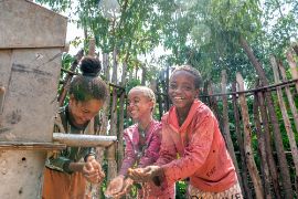 Three laughing children are standing by a pipe from which water is flowing, holding their hands under the stream.