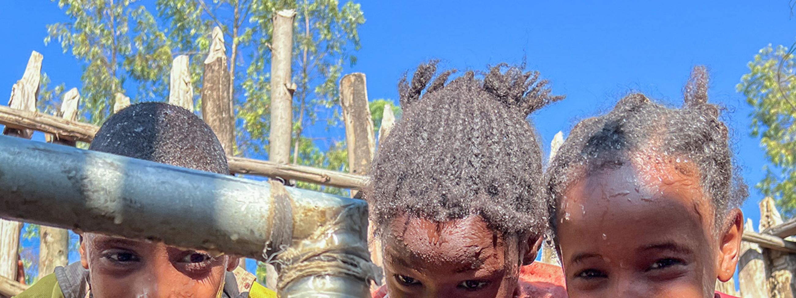 Drei lachende Kinder waschen sich das Gesicht neben dem dicken Wasserstrahl, der aus einem Rohr schießt.