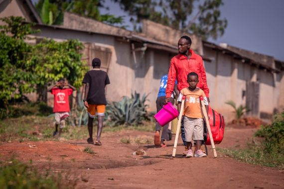 Ein kleiner Junge geht mit Gehstützen einen unbefestigen Weg an Hütten entlang, hinter ihm ein Mann, der eine Tasche und einen Eimer trägt.