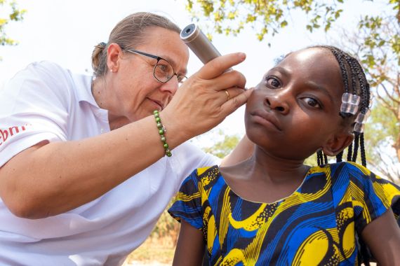 Eine Frau untersucht die Ohren eines etwa neunjährigen Mädchen mit kleinen Zöpfchen und Glasperlen im Haar.