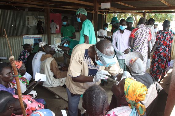 Ein Mann mit Vergrößerungsbrille untersucht die Augen einer Frau, die mit anderen Patienten und Patientinnen auf Holzbänken unter einem Blechdach sitzt.
