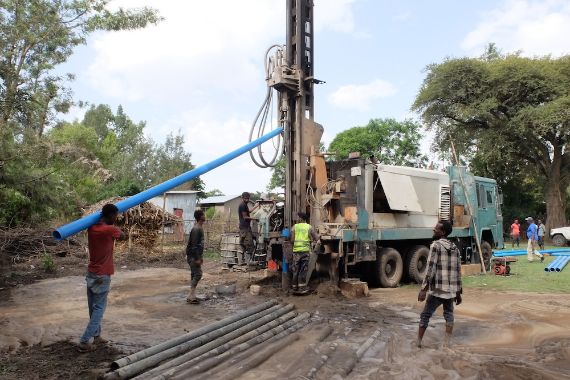 Auf einem Laster steht eine meterhohes Bohrgerät. Männer stehen an dem Gerät, das sich in den Boden schraubt. Auf einem gelben Punkt steht: CBM-Initiative TRACHOMFREI 2030