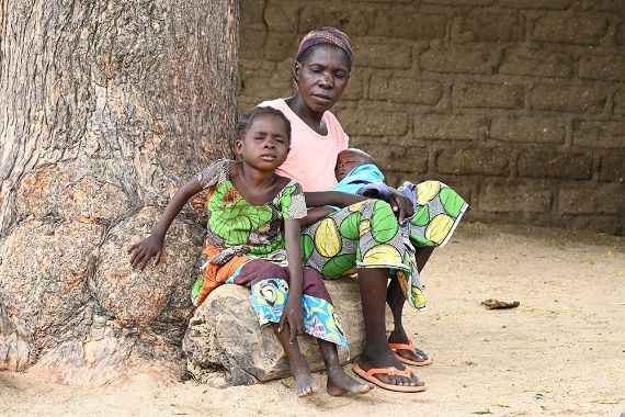 Ein etwa achtjähriges Mädchen und eine Frau mit einem Baby auf dem Schoß sitzen auf einem Holzstück an einem dicken Baumstamm vor einer Mauer.