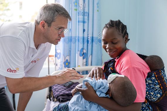Ein Mann im weißen CBM-Polohemd schaut auf den Augenverband eines Kleinkinds, das auf dem Schoß einer Frau sitzt. Im Tragetuch auf dem Rücken der Frau ist ein Baby.