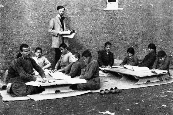 Ein Mann steht neben neun um zwei niedrige Tische auf dem Boden sitzende Jungen. Auf den Tischen liegen aufgeschlagene Bücher, in denen die Kinder mit den Fingern tastend lesen.