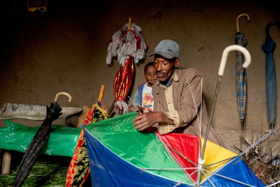A man sitting indoors and fixing an umbrella, a child stands behind him watching. 