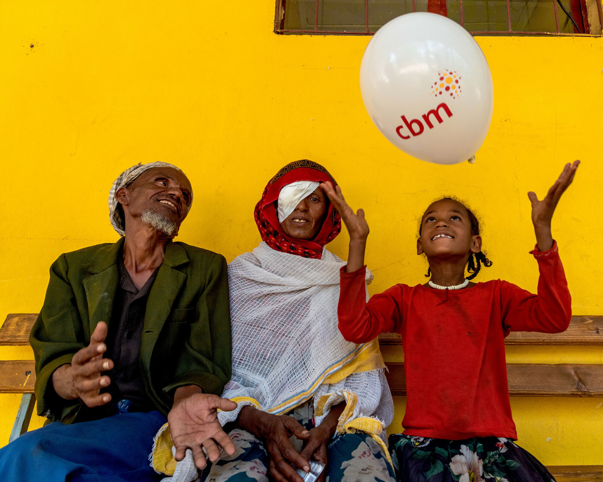 A man, a woman and a child sitting together. The child plays with a balloon, and the woman wears an eye patch over her left eye. 