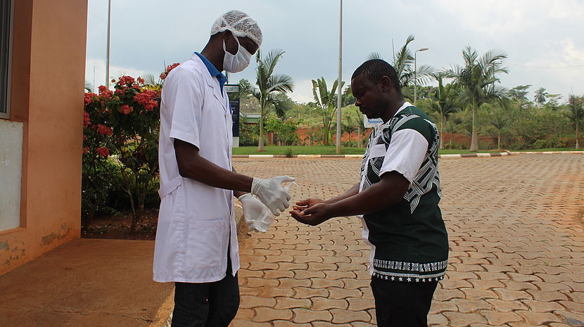 This photo shows two men, one wearing protective gear (face mask, hand gloves and a doctor's coat). He is offering hand sanitizer to the second man, at the gates of the clinic.