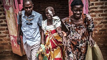 This photo shows an old Ugandan woman with an eye patch being helped by 2 people (a man and a woman)