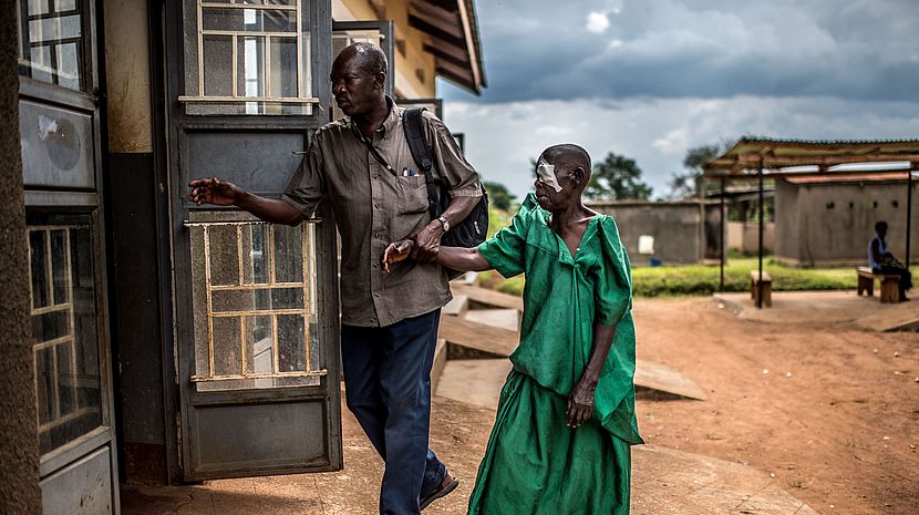 An Ugandan man leads an older Ugandan woman by the hand to a room. One of her eye has a bandage over it.
