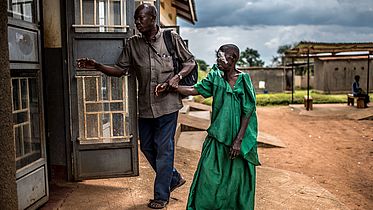 An Ugandan man leads an older Ugandan woman by the hand to a room. One of her eye has a bandage over it.