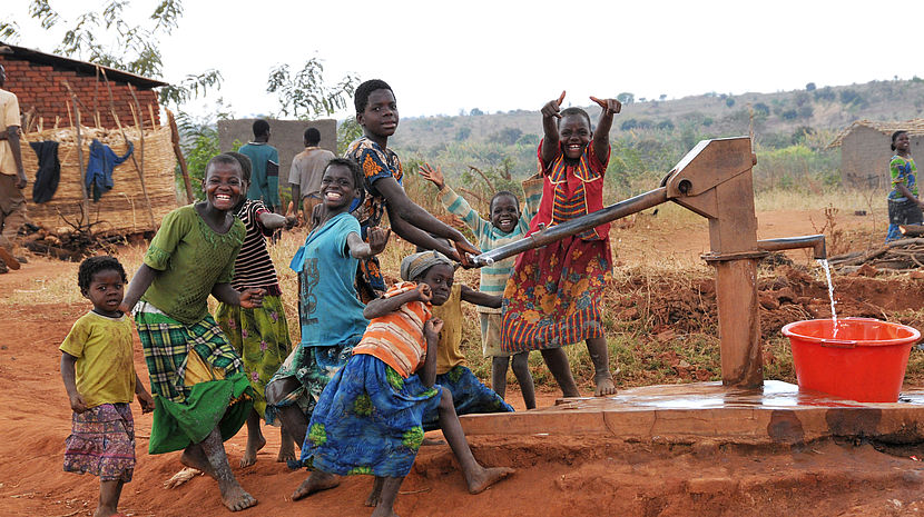 This photo shows a group of children at a water tap community point smiling and laughing together.