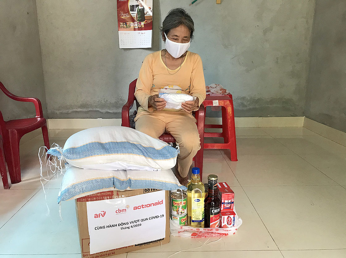 This photo shows a woman in a wheelchair wearing a mask and surrounded by relief material like food and hygiene items.