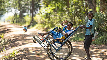 An image showing two young African children on a sunny dirt road surrounded by trees. The child in the foreground is pushing a wheelchair where the other child is seated, waving and smiling at the camera. Both are wearing school uniforms, with the child in the wheelchair in a blue dress and the one pushing in a teal shirt.