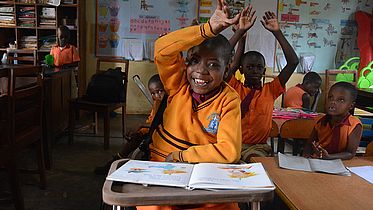 This photo shows a young Ugandan girl in her school, sitting at her desk with her books open. She has her hand raised high and is smiling.
