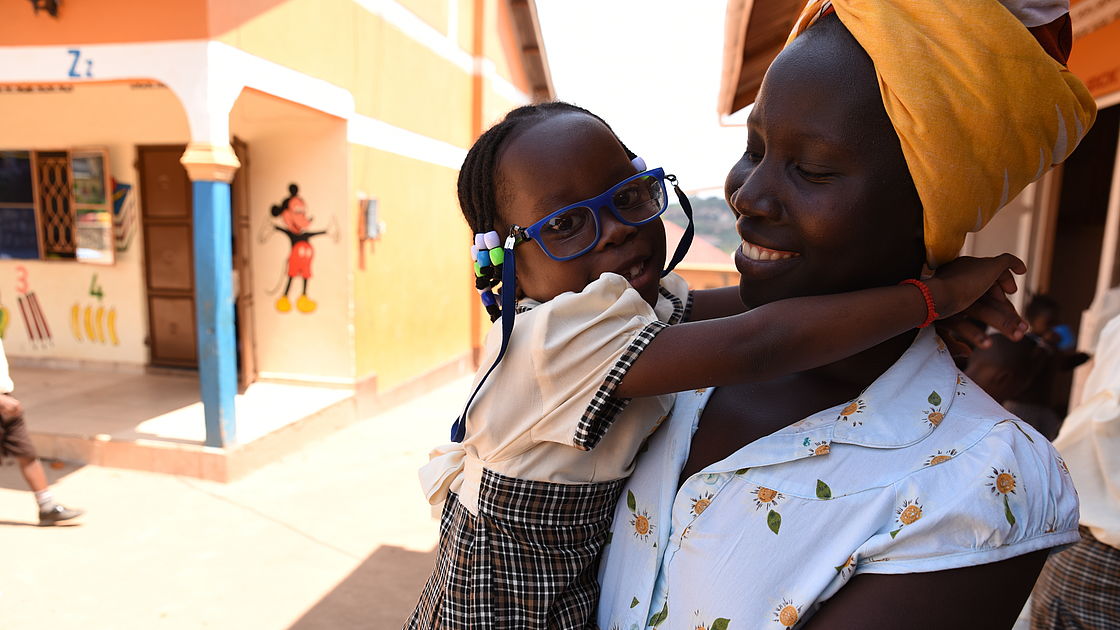 This photo shows a Ugandan woman wearing a headband holding her young daughter in her arms. Her daughter is wearing spectacles and has her arms around her mother.