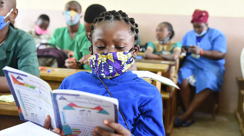 This photo shows a young 11 year old girl studying her notes in her classroom. She is wearing a mask and is surrounded by other students in the background.