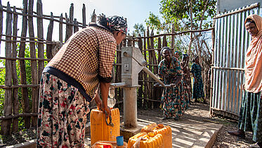 The image shows a group of women at a water well in a rural setting. One woman is actively pumping water from the well into yellow jerry cans, commonly used for carrying water in many parts of the world. Another woman is waiting with her own cans. The scene is outdoors, and it appears to be a sunny day. There's a fence made of sticks and a metal gate in the background, hinting at a simple, communal living environment. The women are dressed in traditional cultural attires.