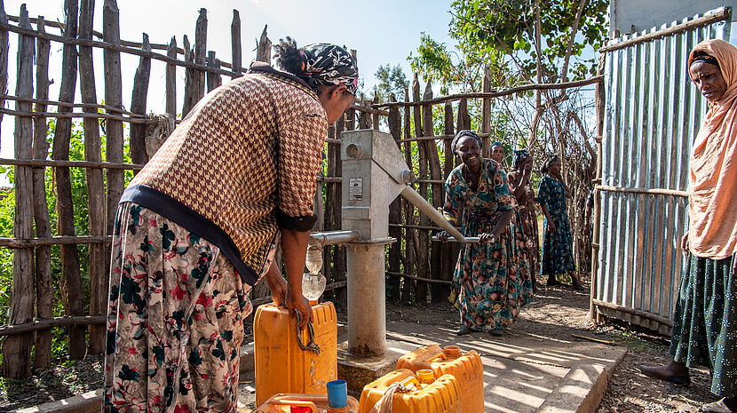 The image shows a group of women at a water well in a rural setting. One woman is actively pumping water from the well into yellow jerry cans, commonly used for carrying water in many parts of the world. Another woman is waiting with her own cans. The scene is outdoors, and it appears to be a sunny day. There's a fence made of sticks and a metal gate in the background, hinting at a simple, communal living environment. The women are dressed in traditional cultural attires.