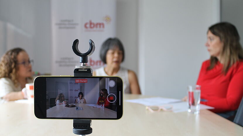 An interview in progress is captured on a smartphone mounted on a tripod, with the recording screen visible showing a time stamp of 00:14:42. Three women are seated at a table, two of whom are blurred in the background with CBM branded banners behind them, while the focus remains on the smartphone and its screen in the foreground.