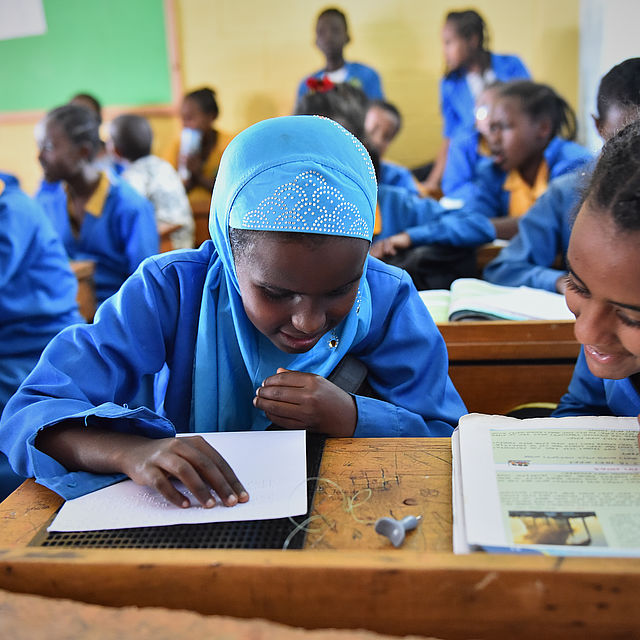 An 11 year old Ethiopian girl with a head scarf wearing a blue school uniform and reading a book in Braille, more students are in the background.