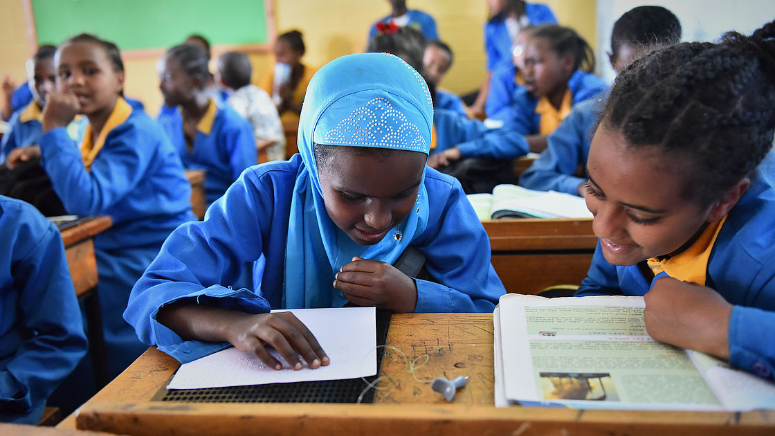 An 11 year old Ethiopian girl with a head scarf wearing a blue school uniform and reading a book in Braille, more students are in the background.