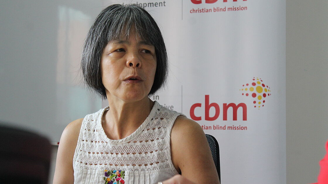 A woman with short grey hair wearing a white sleeveless top with a colorful embroidered design is speaking during an interview. She looks thoughtfully to the side, and in the background, banners with the CBM logo, standing for Christian Blind Mission, are displayed, indicating the setting is an office.