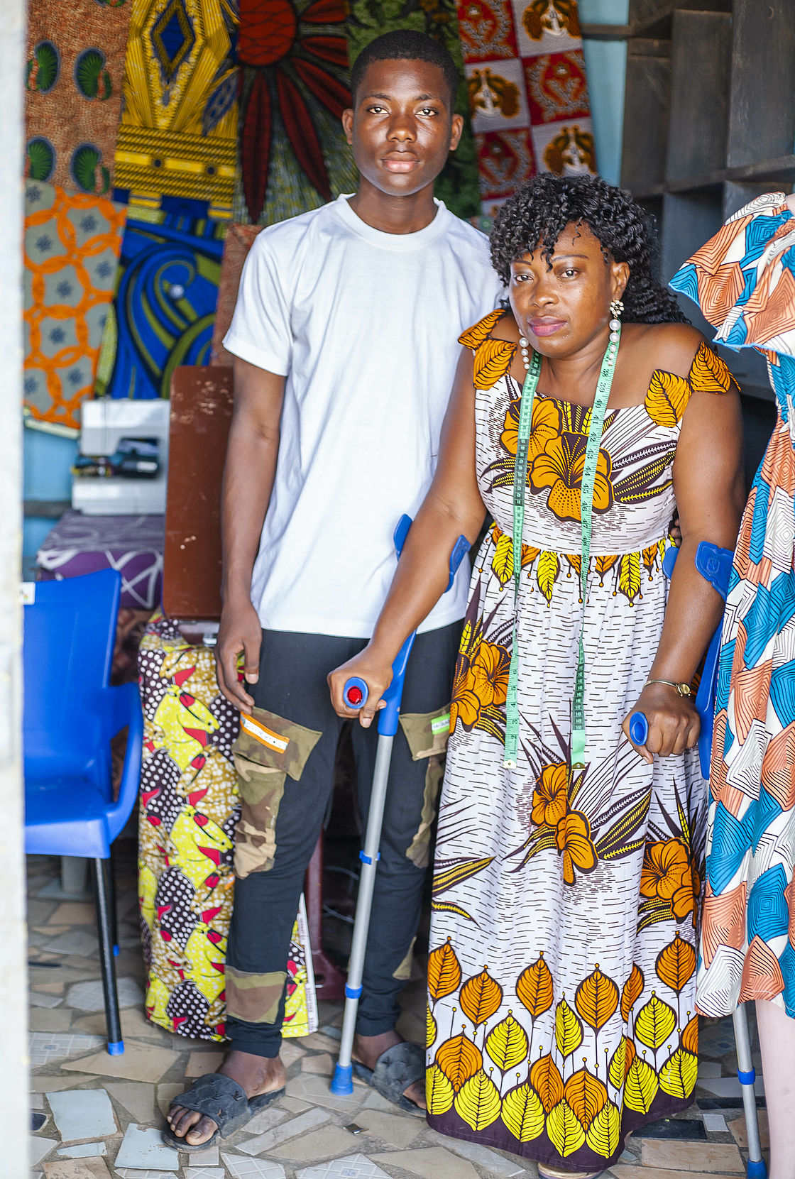 A woman standing with the support of walking stick is standing next to a young man, her son. Around them is colourful cloth in her shop