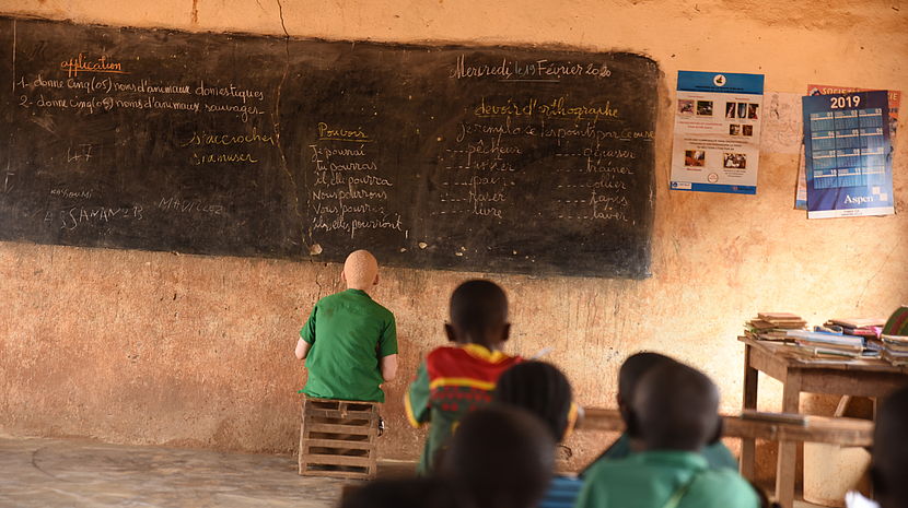 This image shows Assan sitting just a few inches away from the bloackboard in his classroom. He is sitting on a kitchen stool and copying the notes from the board. 